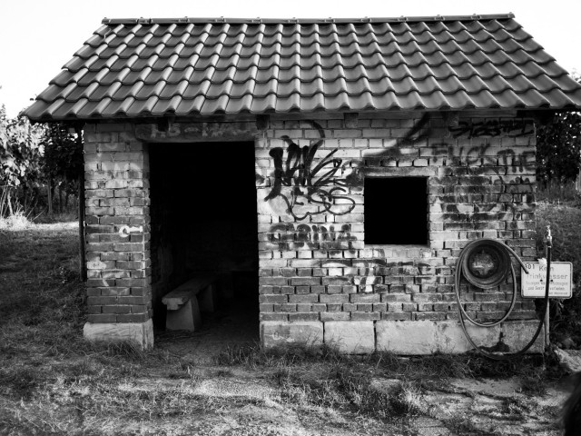 Ein kleines, verwittertes Backsteingebäude mit Ziegeldach. Die Wände sind mit Graffiti bedeckt. Durch den offenen Eingang und das kleine quadratische Fenster ist nur Schwarz zu sehen. Draußen ist ein Schlauch neben einem Wasserhahn aufgewickelt. Schwarz/Weiss Fotografie.
