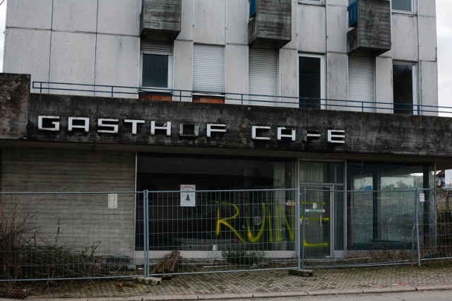 Verlassenes Gebäude mit "Gasthof Café"-Aufschrift und einem Graffiti aus dem Wort „Ruine“, davor ist ein Baustellenzaun.