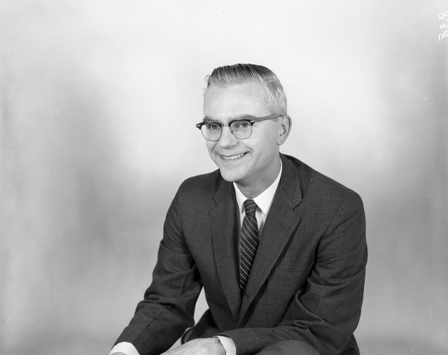 A black and white photo of Frank Drake. He has short hair and glasses, and is wearing a dark suit with a dark, striped tie. Drake is seated, with his left elbow resting on his thigh. He is visible from roughly the waist up. Drake is smiling and looking just to the left of the photographer.
