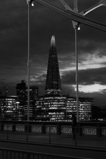 Monochrome photo of The Shard, against a cloudy late evening sky, viewed through the space between two suspender cables of a bridge. The Shard is a stabby looking skyscraper, with the top few levels lit from within. At its foot are more regular, boxy glass buildings, all with most floors fully lit up. In the dim foreground, a road then a metal fence, then a pavement and the iron wall at the edge of the deck of the bridge.