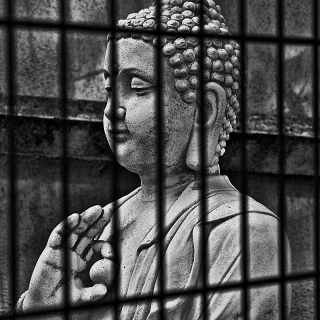 Black and white outdoor photography, close up of a Buddha-like statue behind bars.