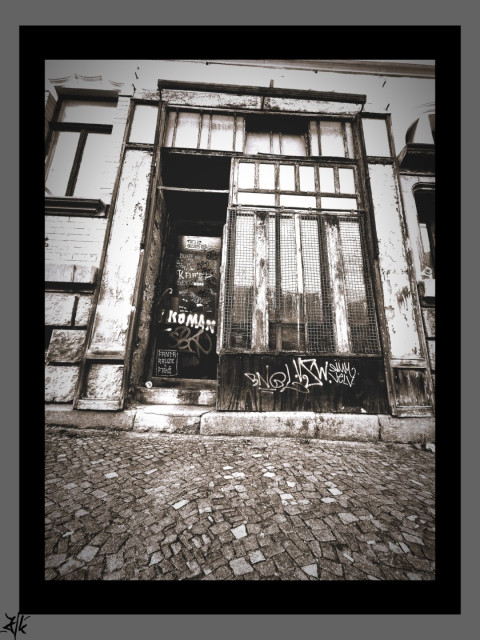 Schwarz-Weiß Foto von einer Hausfront mit einem kleinen schon lange geschlossenem Geschäft.
Es gibt ein kleines Schaufenster aus drei unterteilten Glasteilen und einer Tür die etwas eingelassen ist und mit einer Stufe zu erreichen ist. Die Farbe ist vom Holz schon längst abgeblättert und einige Graffitis zieren jetzt Tür und Fenster.