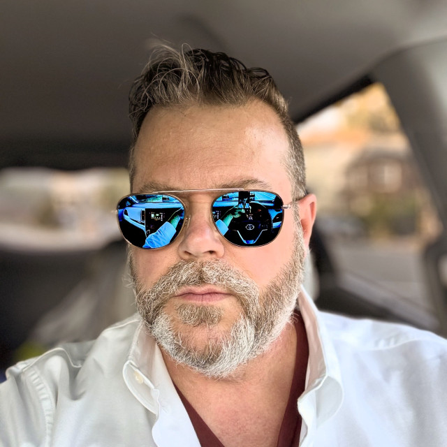 A white guy with brown/blond hair, blue mirrored sunglasses, and a brown/grey beard. He’s wearing a white oxford button-down over a maroon v-neck t-shirt. He looks grumpy, probably because he knows he never photographs well.