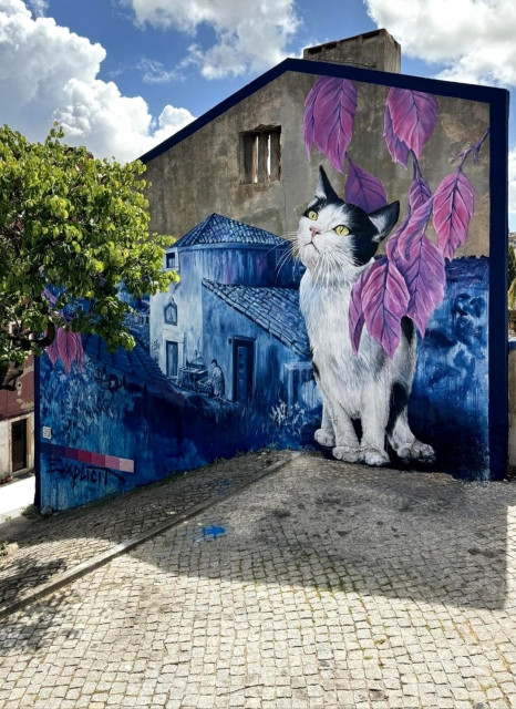 Streetartwall. A beautiful mural with a cat was sprayed/painted on the outside wall of a multi-storey building on a slope. The black and white cat sits under purple leaves and looks upwards. Behind it, the old houses of the village are painted in different shades of blue. An old woman has stepped outside the door and is looking around. The photo shows the unusual paved square (with a tree) in front of the mural, which slopes steeply downwards. The mural thus extends over two floors.