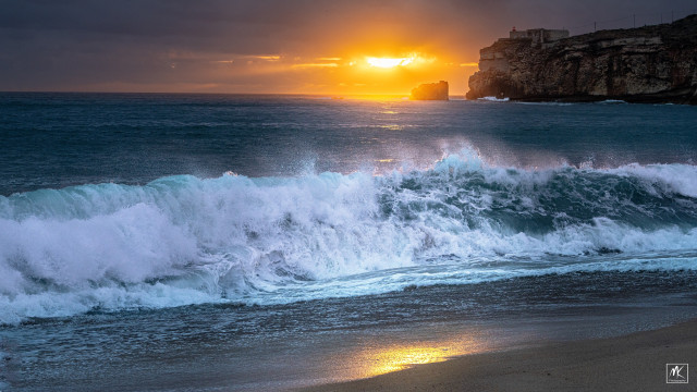 Color photo of a wave breaking on a beach, backlit by the setting sun peeking through clouds near the horizon. In the background to the right is a line of cliffs with a structure and lighthouse at the end and a small rocky islet beyond that, bathed in the glow of the setting sun. 