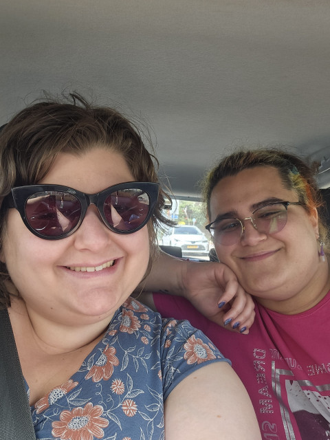my girlfriend and I sitting in a car and taking a selfie.
she's got short wavy hair, and is wearing a blue flowery dress and dark sunglasses.
I have tied coloured hair, and am wearing glasses and a pink shirt.
we're both smiling. 