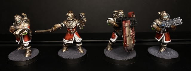 Four painted miniature figures in armor with red-and-white clothing, holding various weapons and a shield, displayed on round bases against a black background.