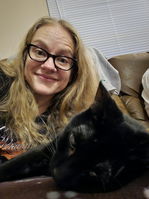Selfie of me leaning over a black cat who us lying on the couch. He looks tired and not impressed but also really cute. I'm smiling because he's so cute.
