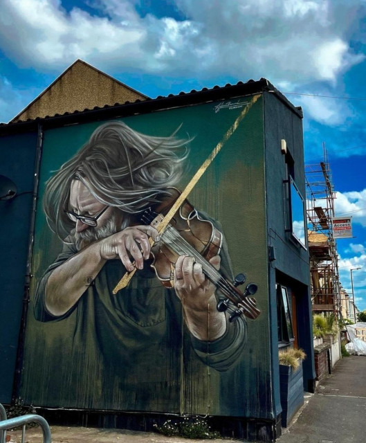 Streetartwall. The portrait of a violinist has been spray-painted on the green exterior wall of a single-storey building. The gentleman with long gray hair, a full beard and glasses is wearing a green T-shirt and passionately playing a violin. The mural is realistically designed and shows him turned away from the viewer, with his hair flying.
Info: The model for the violist was a photo taken by Rupert Engledow (Violin and Veggies).