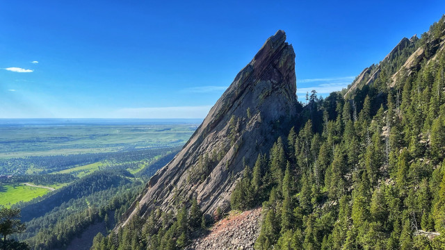 A profile view of the large, sunlit, uptilted Third Flatiron rock sticking above the pine-covered hillside. To the left, an almost endless view of the flat Colorado plains stretching toward the far horizon. Mostly clear skies, just a few thin clouds. 