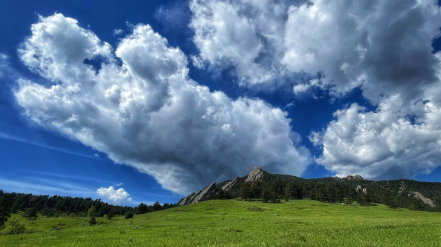 After a long hike, the clouds have changed over the tilted Flatirons rocks, visible in the distance across a rolling, green meadow. There are several, large, dramatic towering cumulus clouds building above and behind the rocks, high into the blue skies. The clouds have darker bases that look somewhat stormy. 