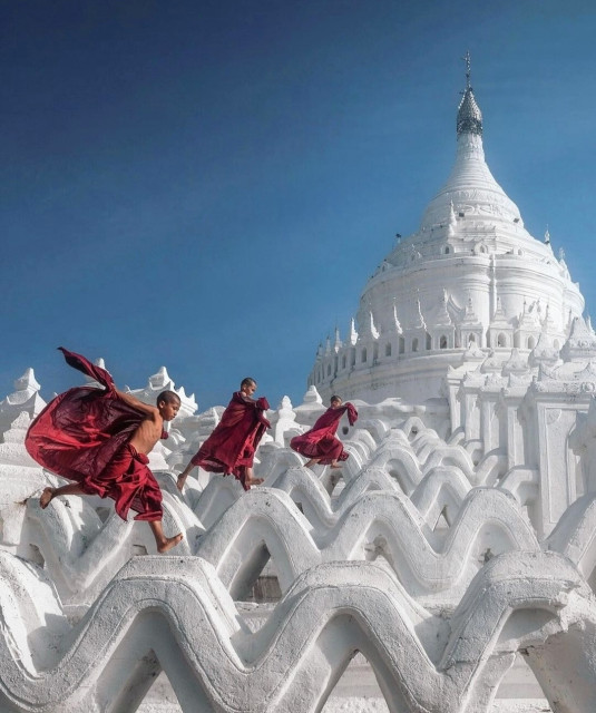 Photography. A color photo of three young monks in red robes jumping over white, wavy parts of a pagoda. The photo shows them in motion and other white parts of the pagoda in the background.
Info: The Hsinbyume Pagoda (also known as Myatheindaw Pagoda) is a Buddhist sanctuary in Mingun (Myanmar). King Bagyidaw had it built in 1816 for his deceased favorite wife Hsinbyume. The pagoda symbolizes the mythical Mount Meru, the center of the world. Seven terraces decorated with waves represent the seven seas which, according to Buddhist belief, surround Mount Meru.