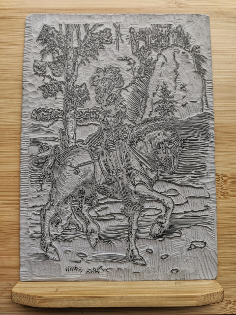 A carved block of gray linoleum depicting Lucas Cranach the Elder's detailed drawing of a knight upon a horse mid trot through a rocky forested landscape with a village nestled in the background below a castle perched upon a stylized mountain.