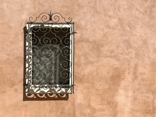 Colour photo of a rectangular window set in a terracotta coloured plaster wall, in direct bright warm sunlight. The window has a white marble frame, and is covered by an ornate metal scrollwork grille, the shadow of which is throwing a strip of curly wavy pattern along the bottom of the window. 