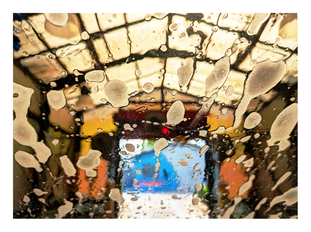daytime. view through a car windshield during a car wash. suds on the glass, skylights and a blue building visible through the exit opening.