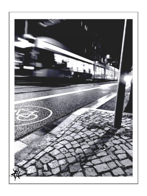 Schwarz-Weiß Foto von einer vorbeifahrenden Straßenbahn bei Nacht. Durch ihre Bewegung verschwimmt ihre Form und die Lichter. Das Foto habe ich von unten gemacht, sehr nahe des Bodens. Vorne ist Kopfsteinpflaster vom Gehweg zu sehen sowie der Radweg auf der Asphaltstraße.