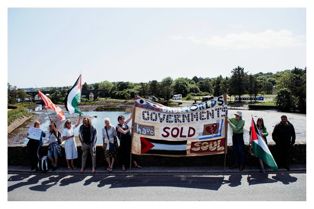 A group of people with Cork and Palestine flags and hand sewn banner in a show of solidarity with the people of Palestine on the Ballydehob bridge with the 12 Arch bridge and estuary in the background.