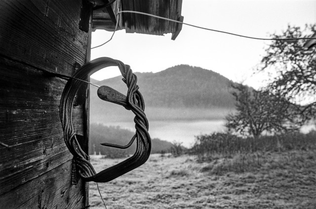 The image is a black and white photo of a loop hanging on the side of a wooden building. The loop is made of intertwined metal wires. It is looped around a metal hook that is screwed into the wood. It appears to be unused.

The building is located in a rural setting in the Carpathian Mountains. There are trees and bushes in the background on the right.

In the background you can see a mountain peak covered with forest. Between the mountain and the building lies a valley covered with fog.