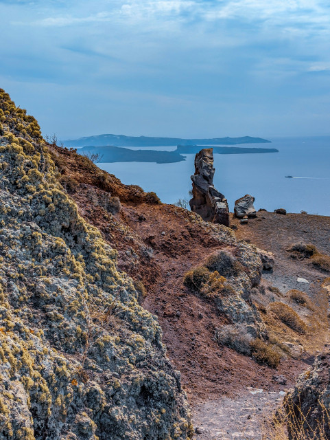 This shot captures the striking volcanic textures of Santorini. 

In the foreground, to the left, there is a prominent lichen-covered outcrop. 

To the centre, a dark, totem-shaped volcanic rock stands against the backdrop of a pale blue ocean and sky. 

To the right, a ship leaves a trail as it sails, with the dark silhouettes of hazy islands on the horizon.