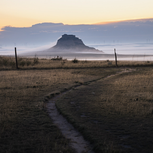 A lone butte is draped in fog in the distance. A dirt trail snakes toward a fence line in the foreground.