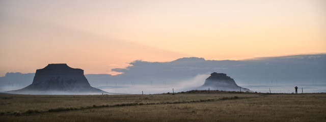 Two buttes at sunrise draped in fog. A lone figure stands in the distance.
