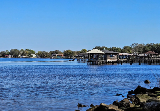 Beneath a sunny, clear blue sky, a view looking out over an inlet of a large blue river, with a rocky shoreline and in the distance a number of boat docks and fishing piers.