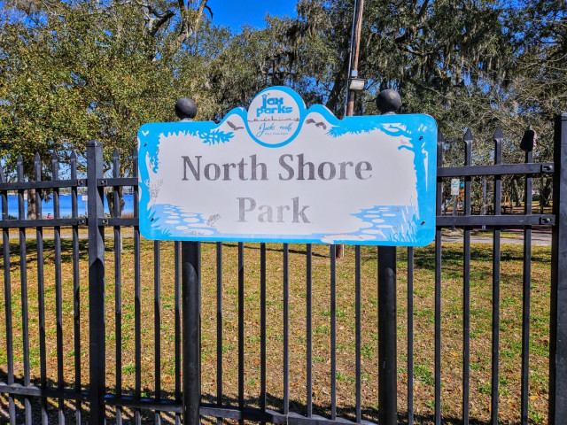 City park sign for Nirth Shore Park, with oak trees, playground equipment, and a partial view of the Trout River in the background.