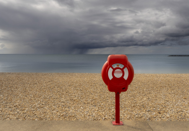 A photo of a seaside scene. A red life preserver holder stands at the edge of a pebble beach. The sun is shining on the beach whilst out at sea a rainstorm rages with heavy rain and black clouds.