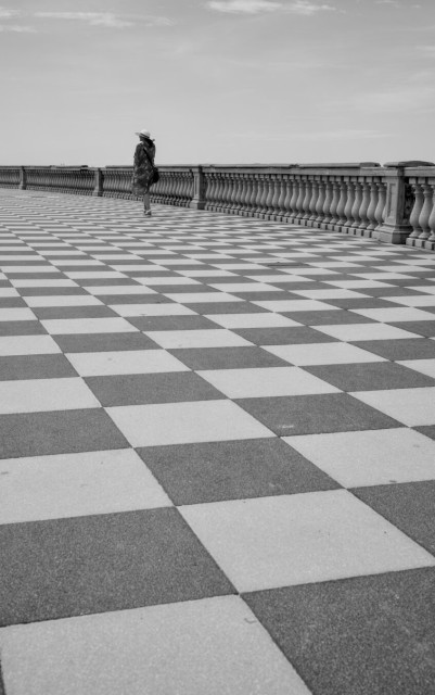 This black and white photograph features a person standing on a large checkered pavement, likely made of alternating dark and light tiles. The scene is spacious and somewhat desolate, with the individual positioned towards the left side of the frame, walking or standing near a balustrade that stretches into the distance. The person is dressed in a hat and a long coat or cloak, creating a sense of isolation or introspection. The sky above is mostly clear with a few scattered clouds. The image captures a sense of vastness and solitude.