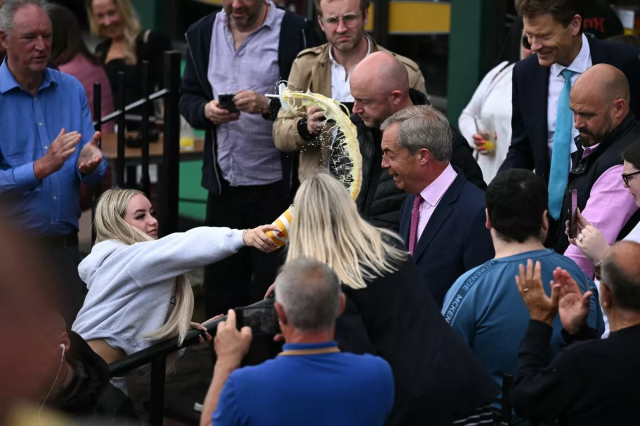 Young blonde woman (probably luring the letch Farage into letting his guard down) throws a yellow milkshake, presumably banana flavoured at a smiling Nigel as his bodyguards do nothing and Richard Tice smiles like a guy who just lost his job to Nigel Farage 

The arc of milkshake is beautiful and righteous 