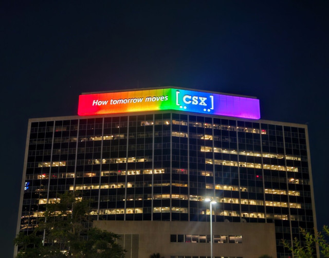 Beneath the dark night sky the large CSX building is illuminated from the countless little office windows with internal lighting glowing bright yellow. The top of the building, with the nameplate, "How Tomorrow Moves: CSX" is illuminated with bright rainbow colors of red, orange, yellow, green,  blue, and purple for the Pride Month of June 2024.