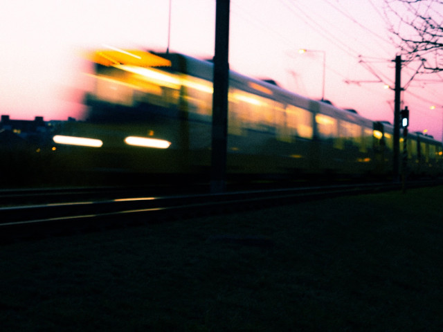 Blurred image of a moving train against a sunset sky with silhouettes of buildings and trees in the background.