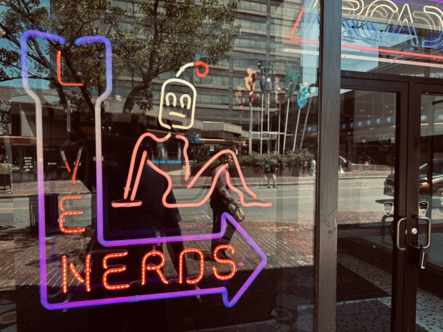 A neon sign of a pin-up style robot sitting on an a L-shaped arrow. Inside the arrow are the words “LIVE NERDS”. This sign is on a bar/arcade in Portland, Maine. 