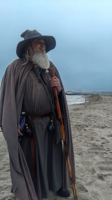middle aged man with grey beard and gandalf style outfit stands smiling on beach in front of Santa Monica pier