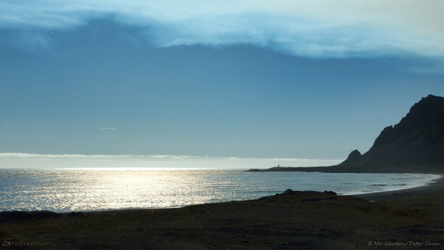 A photo of a landscape. In the foreground is the dark silhouette of a coastline and promontory, and the sea is filling the rest of the bottom third of the shot. The sky is fairly clear cyan except for a few unusual exceptions. The first is at the top of the photo, there is a strange, wispy veil looking like pale blue chiffon. At the very top of the shot, where it becomes more dense, there is a slight yellowish tinge to it. On the horizon, immediately above the sea there is what looks like a slightly opaque layer of thin cloud. This is actually a heavier gas which has settled above the surface of the sea. The water is reflecting sunlight centre left, but it's yellowish and odd.