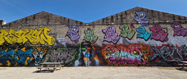 Rear side of a large, double peaked warehouse colorfully decorated with art, graffiti, and murals. Two old, weathered wood picnic tables are still next the abandoned building. Large bubble letters, cartoon characters, a jaguar, and a portrait of a woman wearing sunglasses and a "I ❤️ Dosar" ballcap.