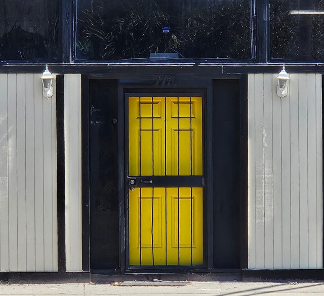 Unusually painted building with the upper portion a long row of darkened out windows, below panel-like siding painted white, with the entrance door a bright yellow with wide black trim and thick black security bars.