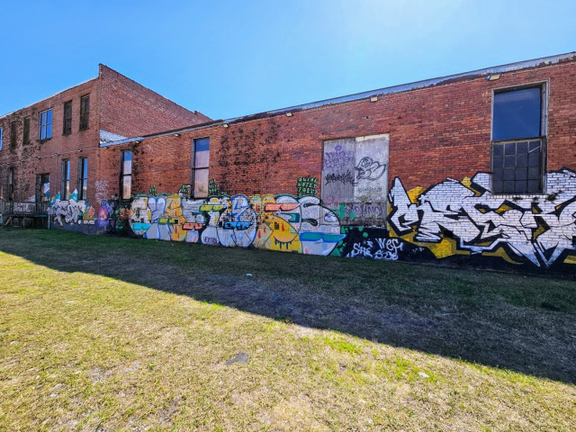 Large red brick industrial building, long out of use, with boarded doors and windows, and vines climbing the exterior walls. Colorfully decorated with large bubble letter graffiti.