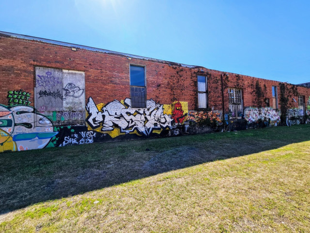Large red brick industrial building, long out of use, with boarded doors and windows, and vines climbing the exterior walls. Colorfully decorated with large bubble letter graffiti. In the center, a red caped Orco character from the 80's cartoon "He Man and the Masters of the Universe"
