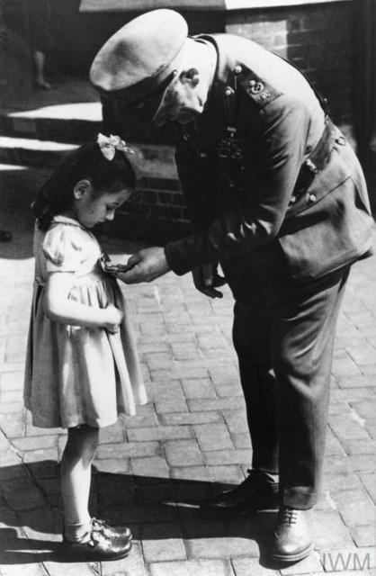 photo of Tania Szabo. She is a five-year old white girl in buckle shoes. A senior Army officer is leaning down to look at the George Cross pinned to her summer dress.