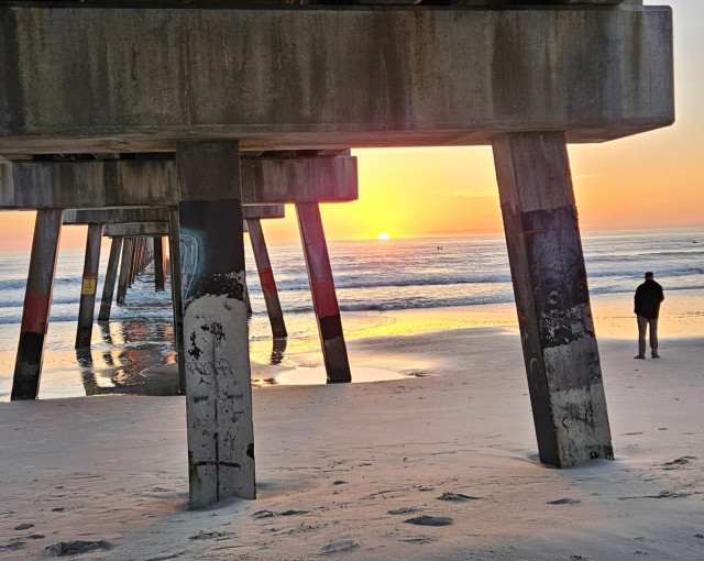 Bright yellow sunrise over the Atlantic's horizon casting shades of orange, yellow, and red across the sky, and reflecting upon the wet sand on the beachfront from beneath an enormous pier.