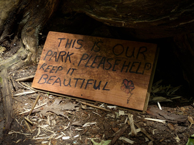 A hand made wooden sign sits in the hollow base of an old tree. The sign reads "This Is Our Park. Please Help Keep It Beautiful" and a small red flower is also drawn on.
