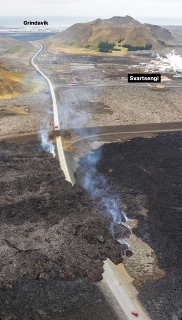 A drone view of the main road into Grindavík. In front of a small red car, then lava covering the road and there is a bit of smoke, then a barrier, to the right in the back  is Svartsengi and in the very back Grindavík.