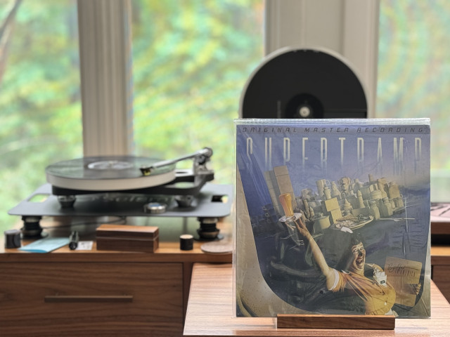 Supertramp - Breakfast in America LP cover.

Out a plane window, a giant waitress replaces the Statue of Liberty, holding a large glass of orange juice on a plate as the torch. As the boot, a menu. An all white city is in the background.

The black LP plays on a Rega NAIA turntable to the left.