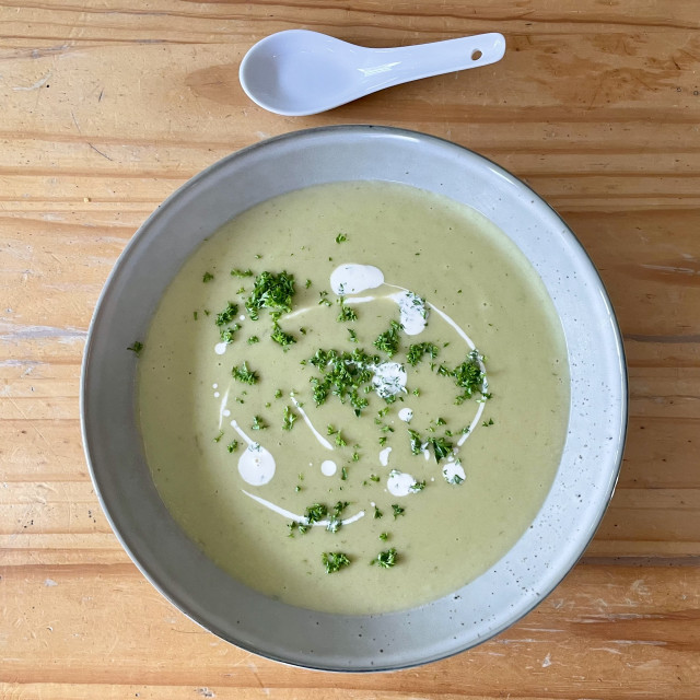 A bowl of greenish creamy looking soup in a shallow bowl garnished with vegan cream and parsley. Overhead, from the top on a timber table. 