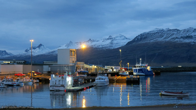 A photo of a landscape, with snow-capped mountains on the horizon. The light is fading but it's still possible to see that the sky is overcast. In the foreground is a compact but busy harbour, with a range of watercraft tied up, sized from row-boats to quite large fishing vessels. A few containers are visible on the dockside, as well as what appears to be a control tower, possibly the harbour-master. Overhead lights are casting pools of yellow light across the vessels and continuing towards the viewpoint across the water in the foreground of the shot.