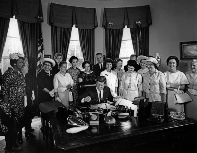 John F Kennedy seated in the Oval office, holding a pen. Thirteen women are standing around him. Twelve are white, and one is black. Someone must have told a good joke as everyone is smiling or laughing.