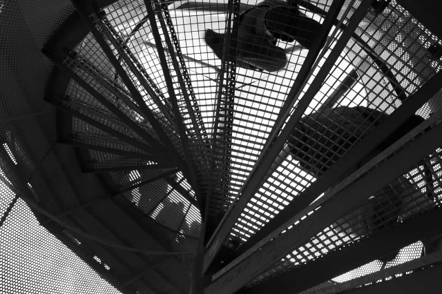 Das Foto zeigt den Blick von unten nach oben aus der Froschperspektive durch die vergitterten Trittstufen einer Wendeltreppe, die auf einen Aussichtsturm führt. Mehrere Menschen sind von unten teils auf der Treppe, teils am Geländer des Aussichtsturms zu sehen.