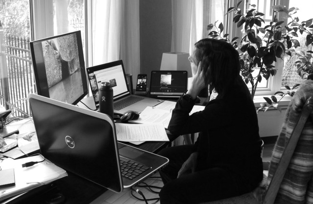 Black and white photograph of a woman sitting at desk at home, holding her head with one hand, surrounded by two computers, a large screen, a tablet and a phone, all open and in use. Two windows with white curtains and a big citrus tree are nearby. The woman is clad in black and a striped wool blanket is on the back of the chair. Quite disconnected from everything at that moment, despite being too well connected to the rest of the world with five screens.