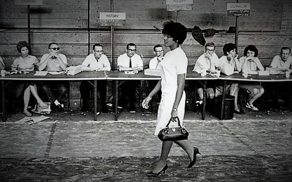 Vivian Malone walking through a registration hall. Behind her is a long table with a row of white people watching her. She is a black woman wearing a white dress.